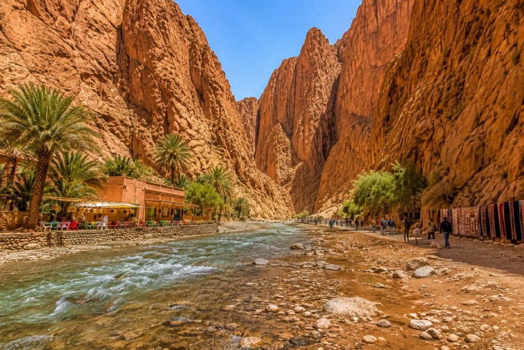 Panoramic view of Todra Gorge in Morocco featuring towering limestone cliffs, the Todra River, and visitors walking through one of the country's most spectacular natural canyons.