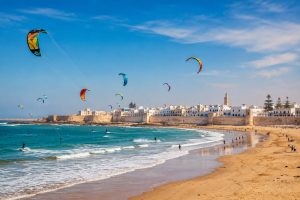 Panoramic view of Essaouira Beach with colorful kitesurfers, golden sandy beach, Atlantic Ocean, historic Medina, and Skala de la Ville in Essaouira, Morocco.