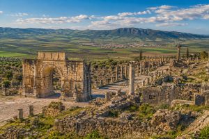 Panoramic view of the ancient Roman ruins of Volubilis featuring the Triumphal Arch, Roman columns, historic streets, and the surrounding countryside near Meknes, Morocco.