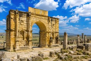 Ancient Roman stone street leading to the Triumphal Arch of Volubilis, surrounded by historic columns and UNESCO archaeological ruins near Meknes, Morocco.